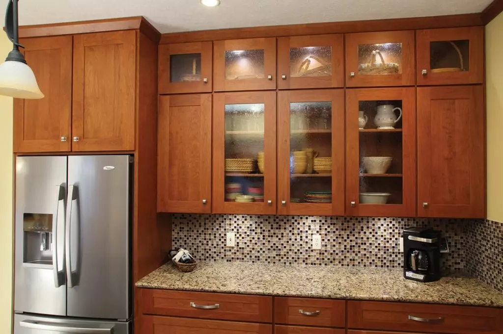 Warm cherry wood refaced kitchen cabinets featuring glass-front upper display doors, a mosaic tile backsplash, and a stainless steel refrigerator.