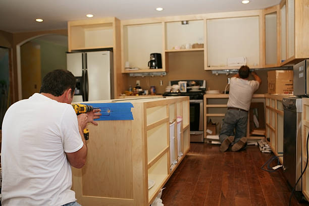 Cabinet maker installing custom made maple cabinets to kitchen island, while electrician installs under-cabinet lighting. 