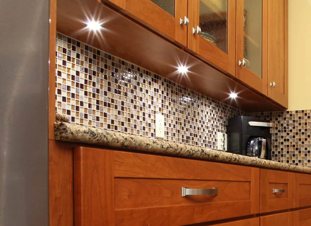 Kitchen cabinet with under-cabinet lighting, featuring light wood doors, drawers, and a dark countertop.