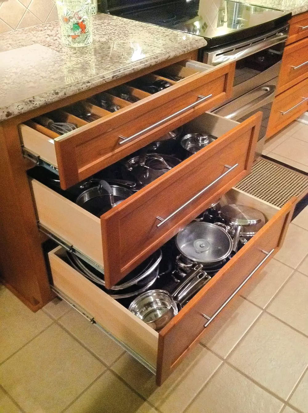 Three open wood kitchen drawers organized with a utensil tray on top and stainless steel pots, pans, and lids in the deeper lower drawers.