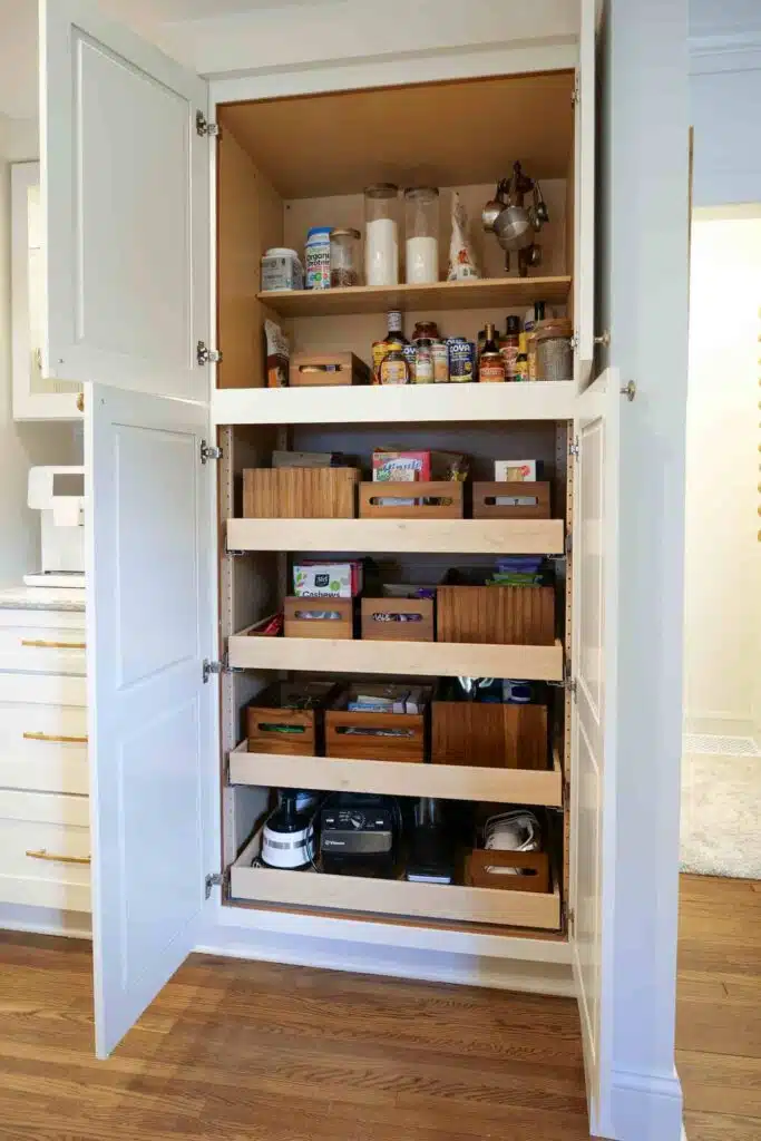 Open white kitchen pantry featuring custom solid wood roll-out shelves neatly filled with organized groceries.
