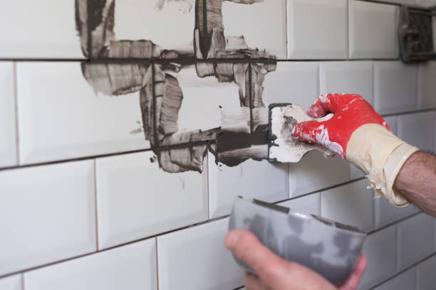 Worker applies grey grout at white tiles in the kitchen backsplash.